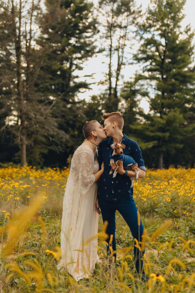 A couple kissing in a field while one holds a dog
