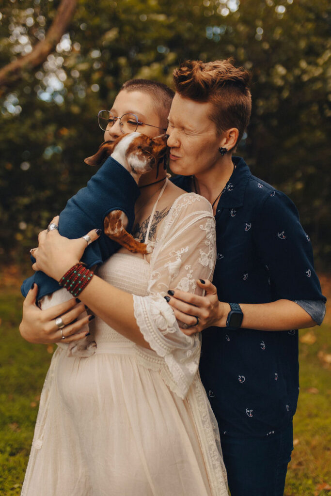A person holding a dog as it kisses the face of another person behind them 
