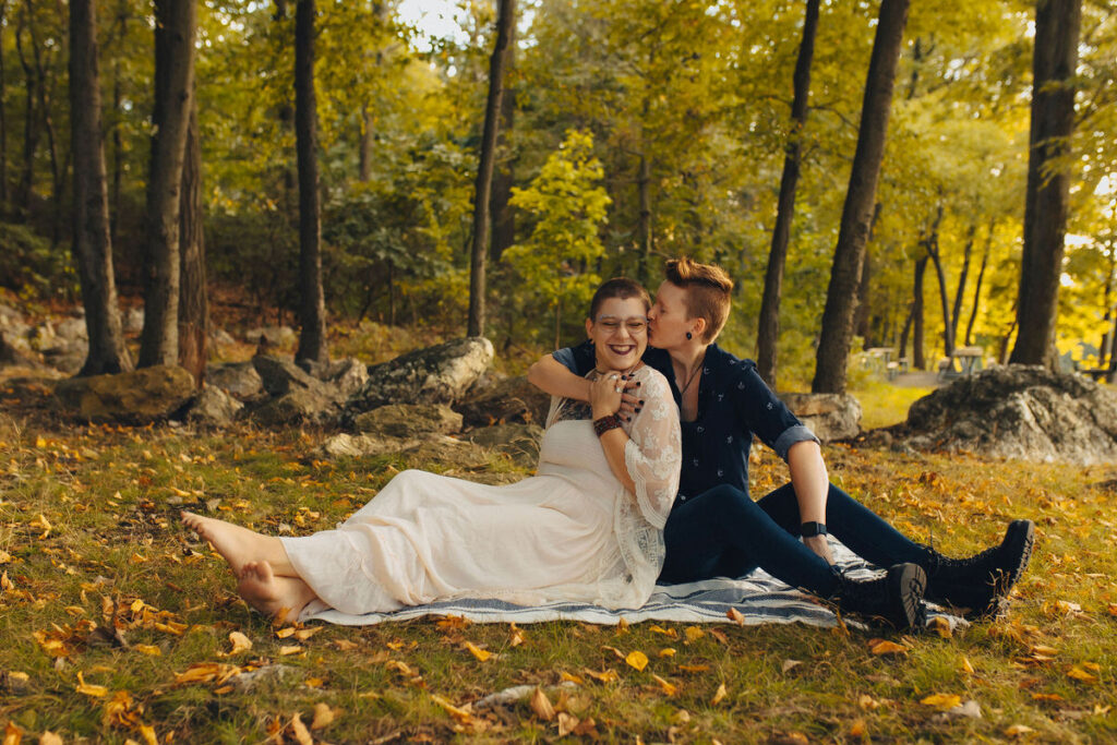A person kissing their partner's cheek as they sit on a blanket in a wooded area 