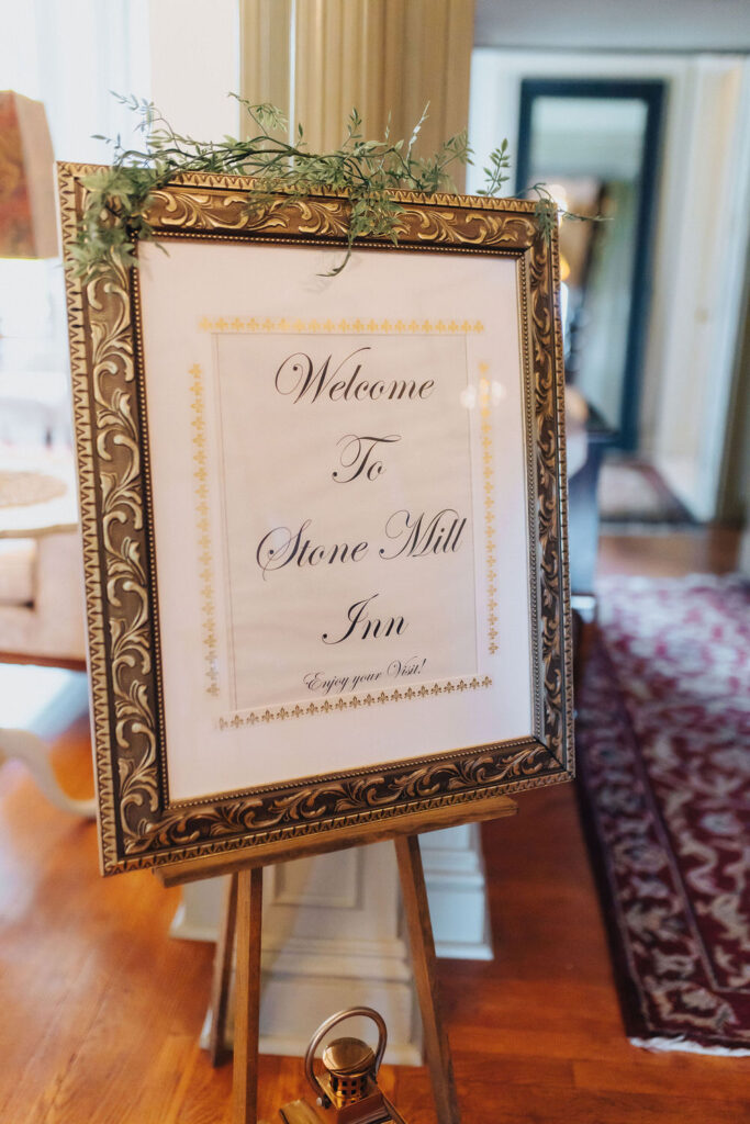 A decorative welcome sign for Stone Mill Inn with ornate framing and a simple, welcoming message for guests, set against an elegant interior background.
