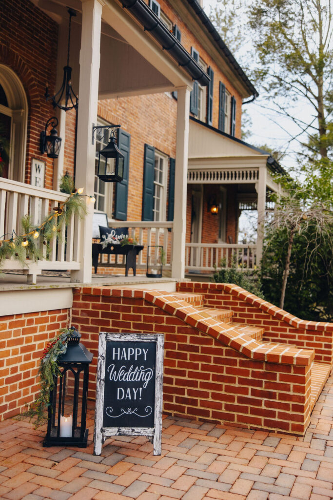 A wedding "Happy Wedding Day!" sign standing in front of a brick-built house with green wreaths and fairy lights.