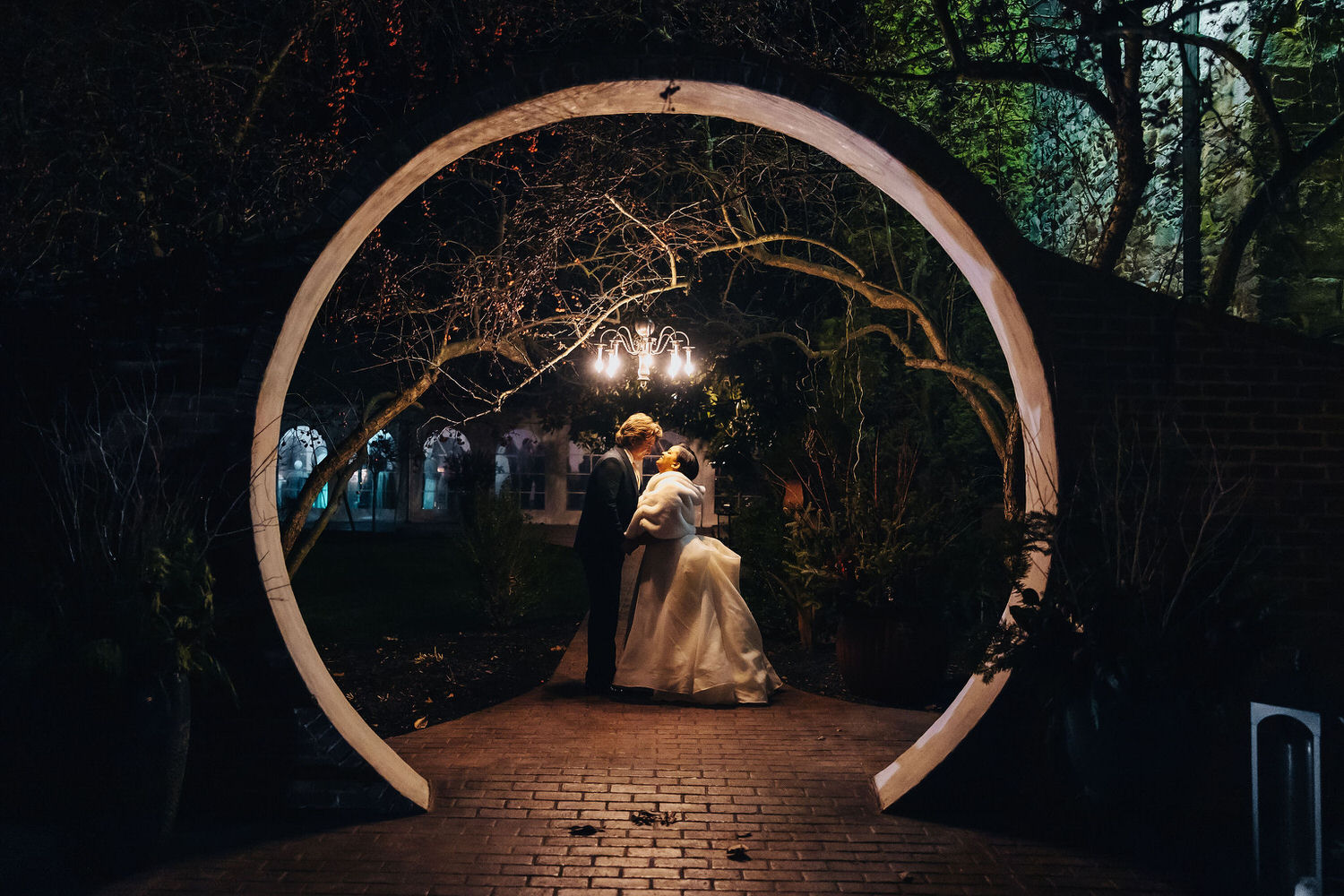 A night-time wedding scene under a large circular archway, showing a bride in a white gown and a groom in a suit embracing each other, with dimly lit trees and a chandelier in the background enhancing the romantic ambiance.