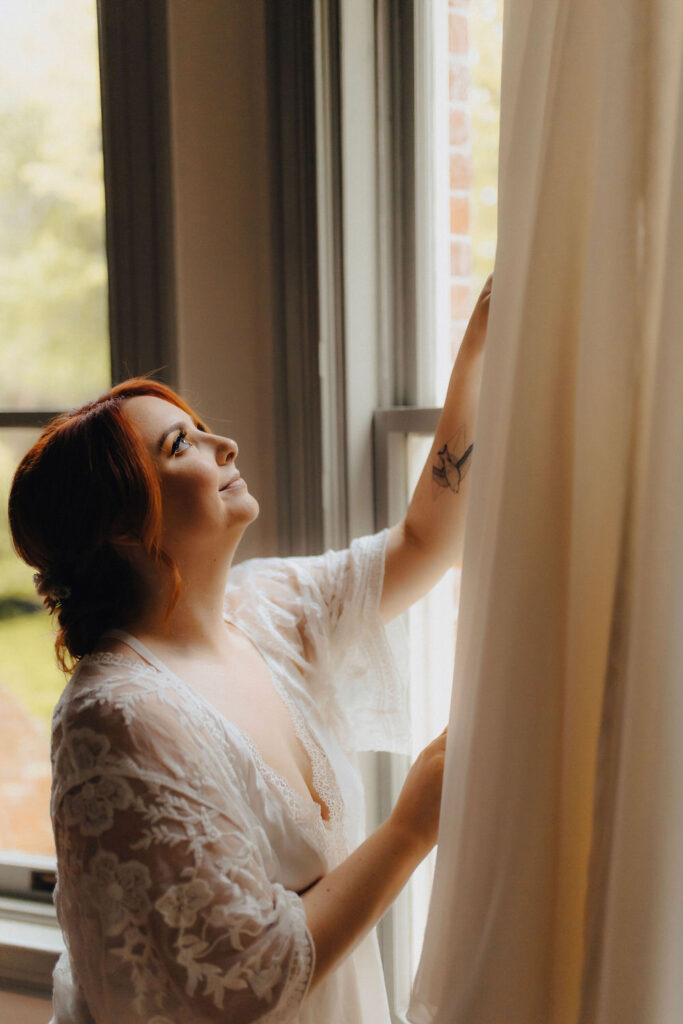 A bride dressed in a light, lace bridal robe looks thoughtfully out a window, her hand gently touching the curtain, in a serene moment before her wedding.
