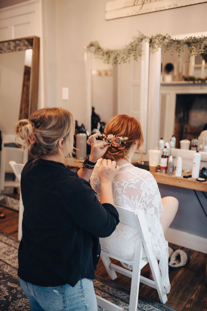 A stylist secures a decorative floral hairpiece in the hair of a bride seated in front of a mirror, reflecting a serene bridal preparation scene in a well-lit, elegant room.