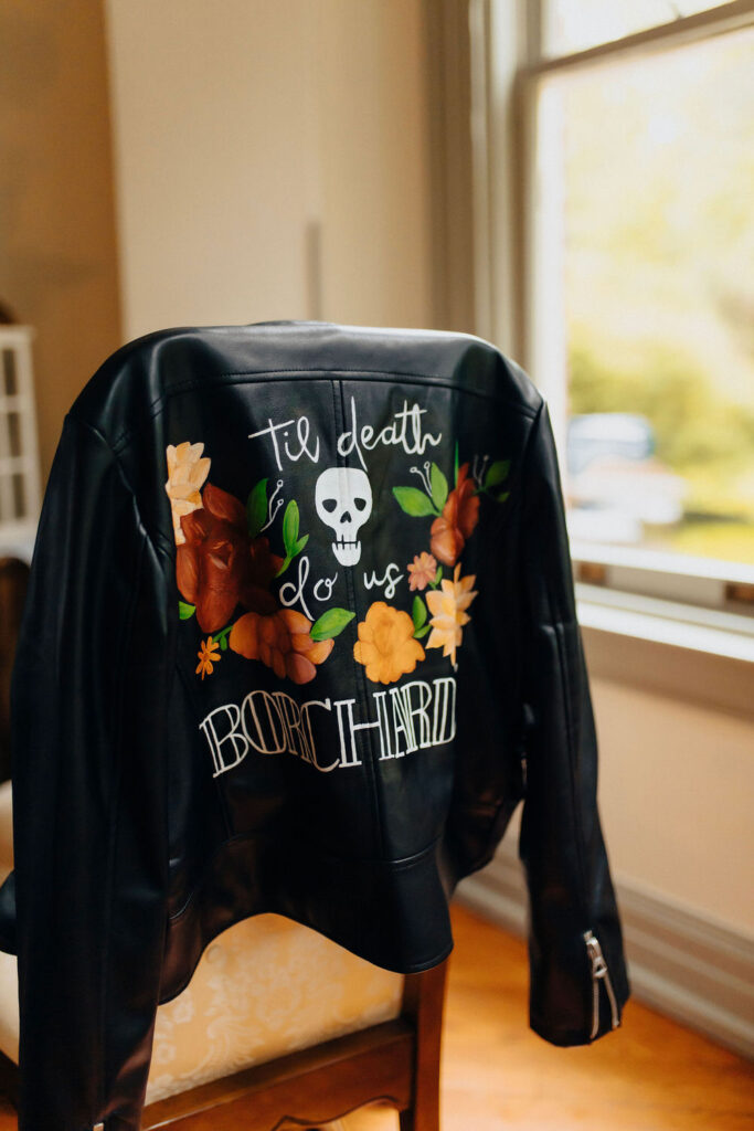 A custom-painted leather jacket hung on a chair, detailed with the phrase "Til death do us Borchard," adorned with a skull and floral design, providing a unique and personalized wedding element.