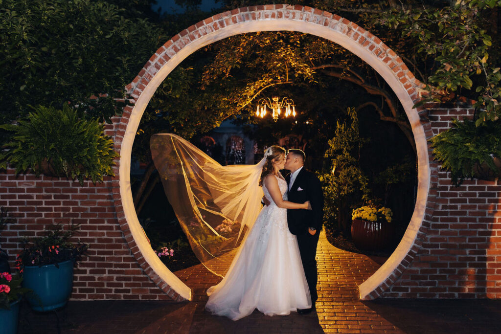 A bride and groom share a kiss under a lit circular archway in a night garden setting, the bride's veil dramatically caught in a breeze, adding movement and a fairy-tale quality to the scene.