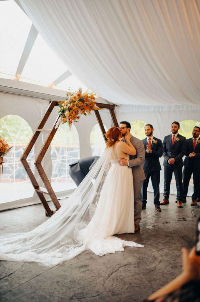 Bride and groom share their first kiss as married couple under a rustic wooden arch decorated with flowers at their tented wedding venue, surrounded by cheering friends and family.
