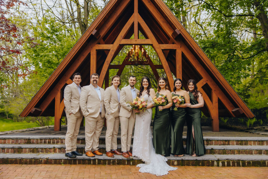 A bridal party under a wooden A-frame outside, featuring the bride in the center flanked by groomsmen and bridesmaids in beige suits and green dresses, respectively, all smiling in a lush green setting.