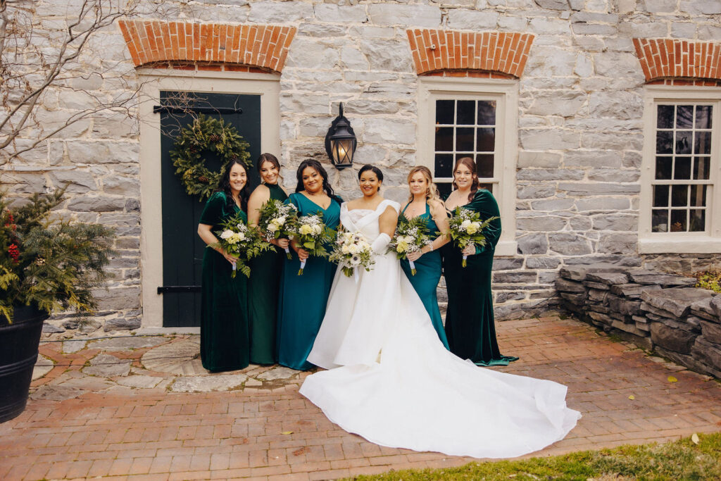 A bridal party posing in front of a historic stone building, the bride in a white gown surrounded by bridesmaids in green dresses, each holding a bouquet, creating a timeless and elegant group portrait.