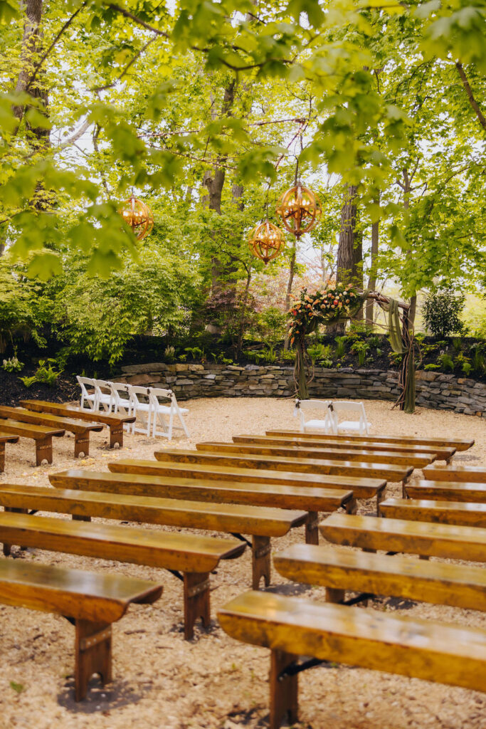 serene outdoor wedding venue setup with rows of wooden benches on sand, surrounded by lush greenery and hanging decorative elements.