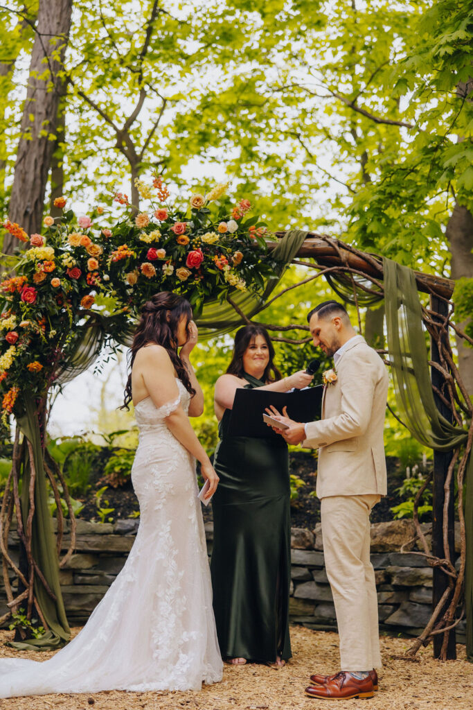 A bride and groom stand under a beautifully adorned floral arch during their wedding ceremony, surrounded by trees, with the groom reading his vows.