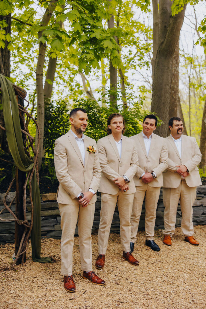 Four groomsmen standing in a line, dressed in beige suits and brown shoes, in an outdoor setting with lush green trees in the background.