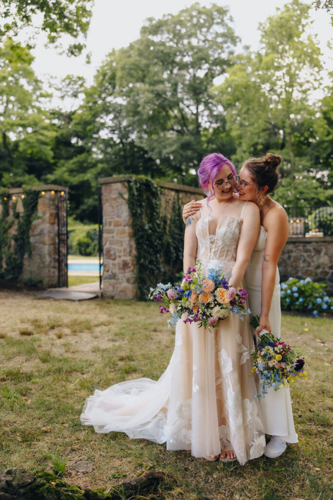 A person standing behind their partner with their head on their shoulder as they both smile. 