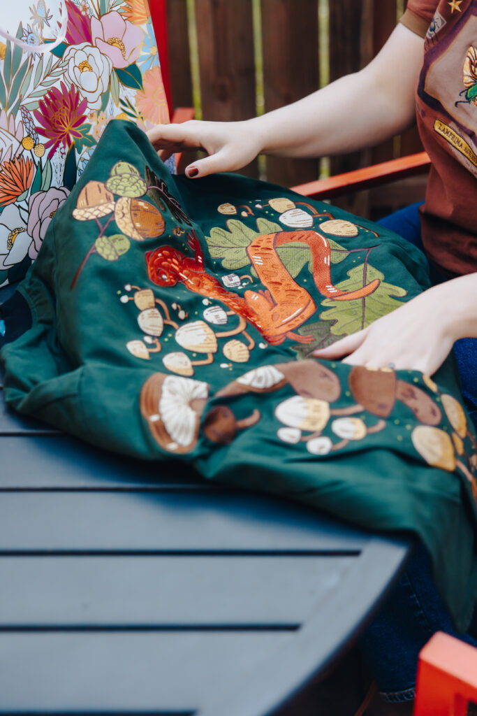 A close-up of a woman's green jacket that has mushrooms and acorns embroidered on the back.