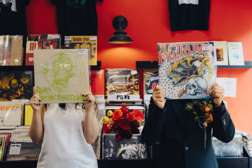 A couple standing side by side, each holding a record in front of their face in a record store with a bouquet or orange and red flowers between them.