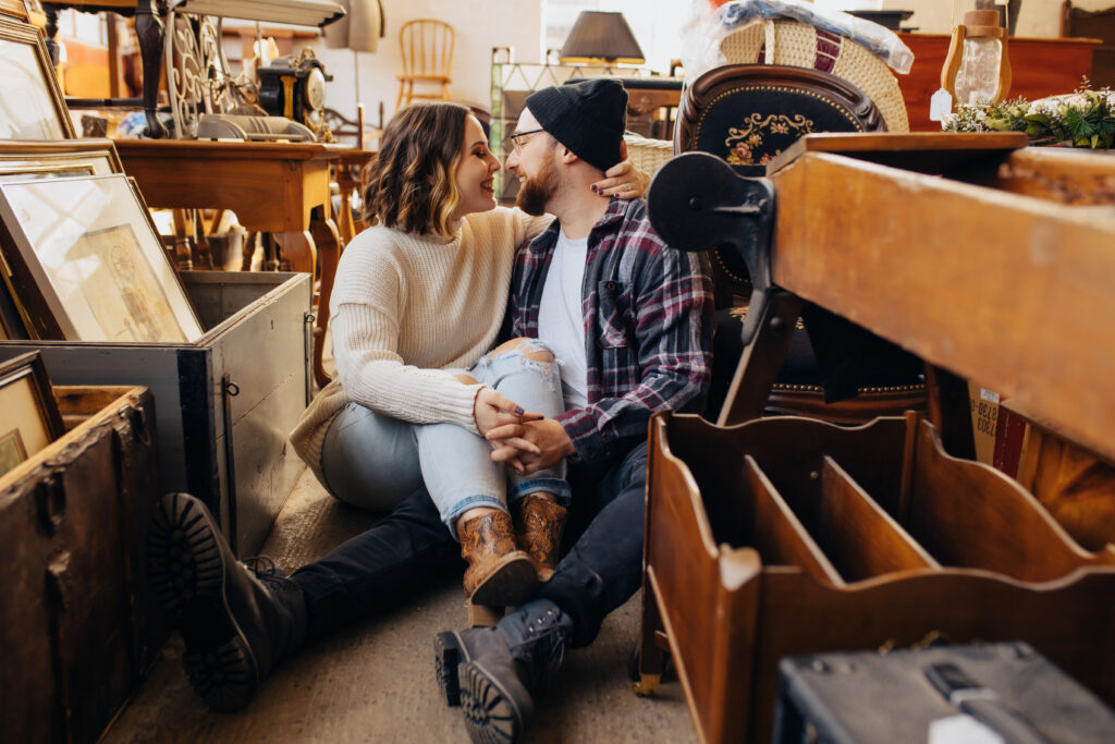 A woman with her legs over her fiance's leg as they sit on the floor and smile at each other
