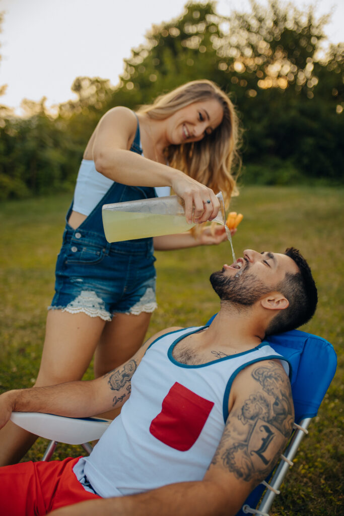 A woman wearing jean overalls, pouring lemonade into her fiance's mouth as he sits in a lawn chair.