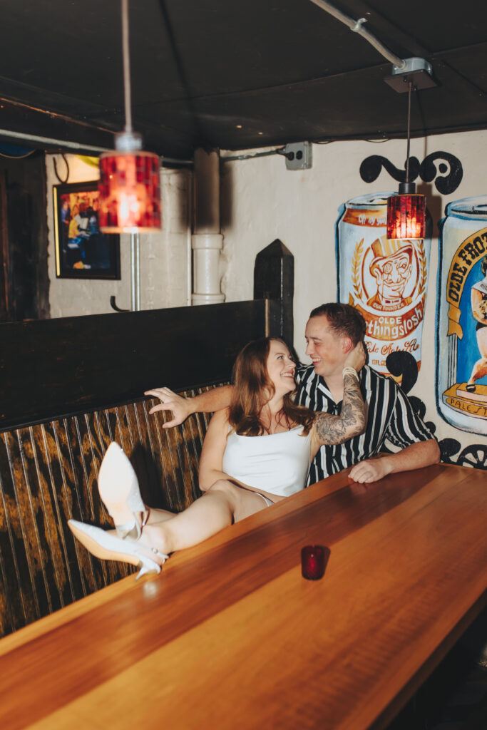 A woman wearing a white dress, leaning against her partner in a dive bar booth.