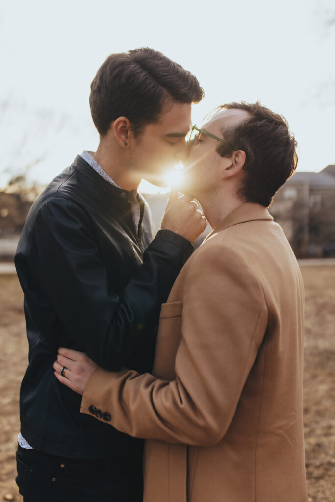 Two men kissing with the sun glowing in the background during their engagement session photoshoot.