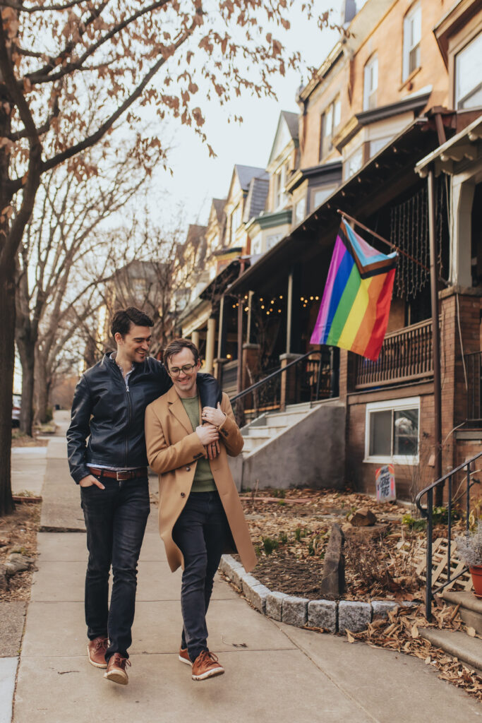 A gay couple walking down the street, with one arm slung over the other's shoulder as they both smile.
