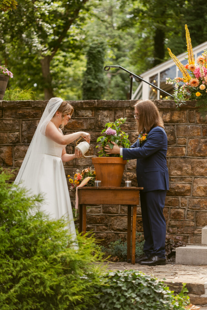A couple in their wedding clothes watering a plant together as an example of a soil unity ritual done during their wedding