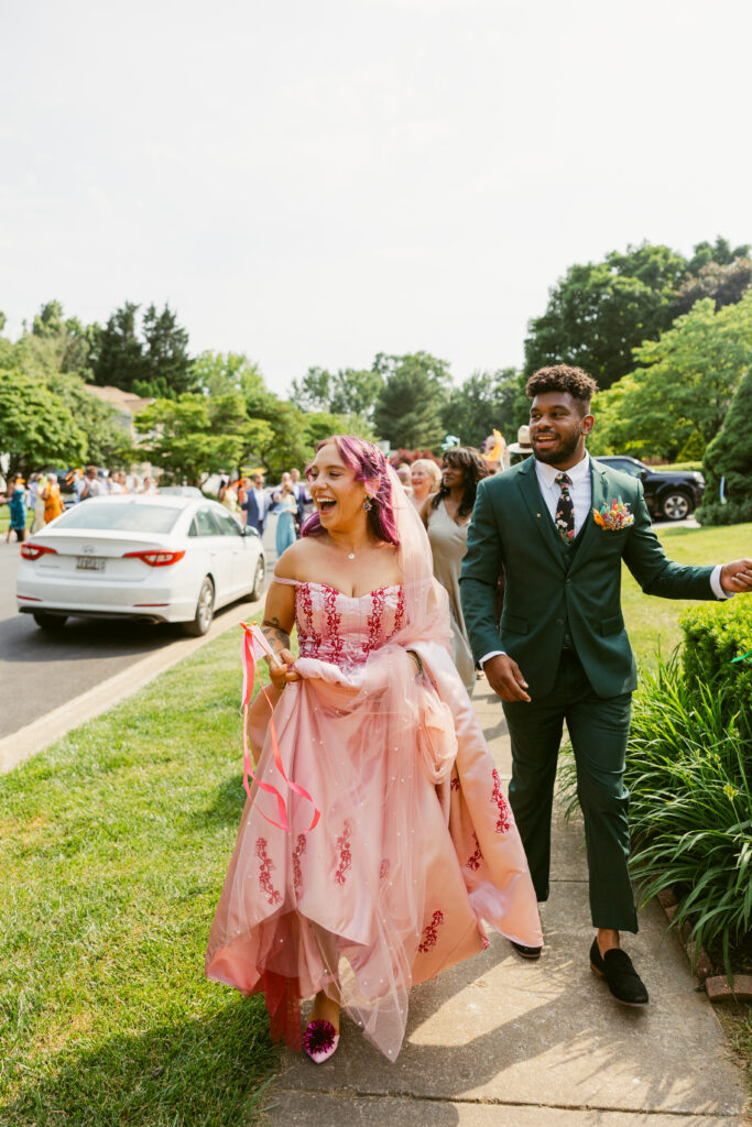 A bride wearing a pink dress with purple hair and a groom wearing a dark green suit, smiling as they walk down a neighborhood street together