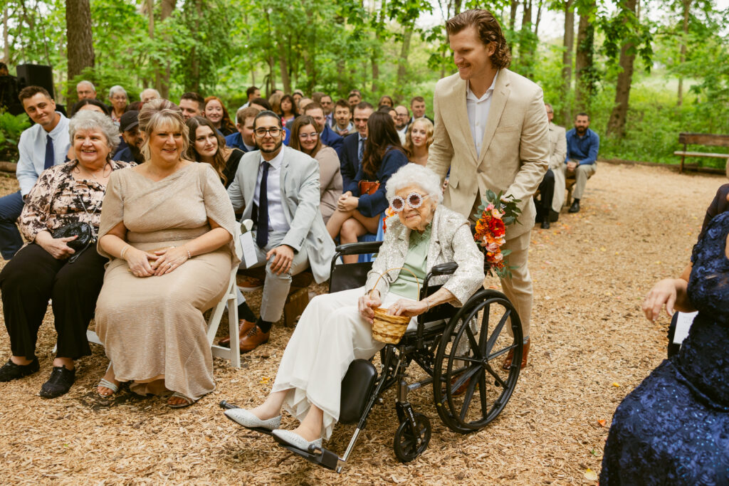 A man wearing a tan suit wheeling a grandma down the wedding aisle. She is wearing bedazzled sunglasses and throwing petals from her basket
