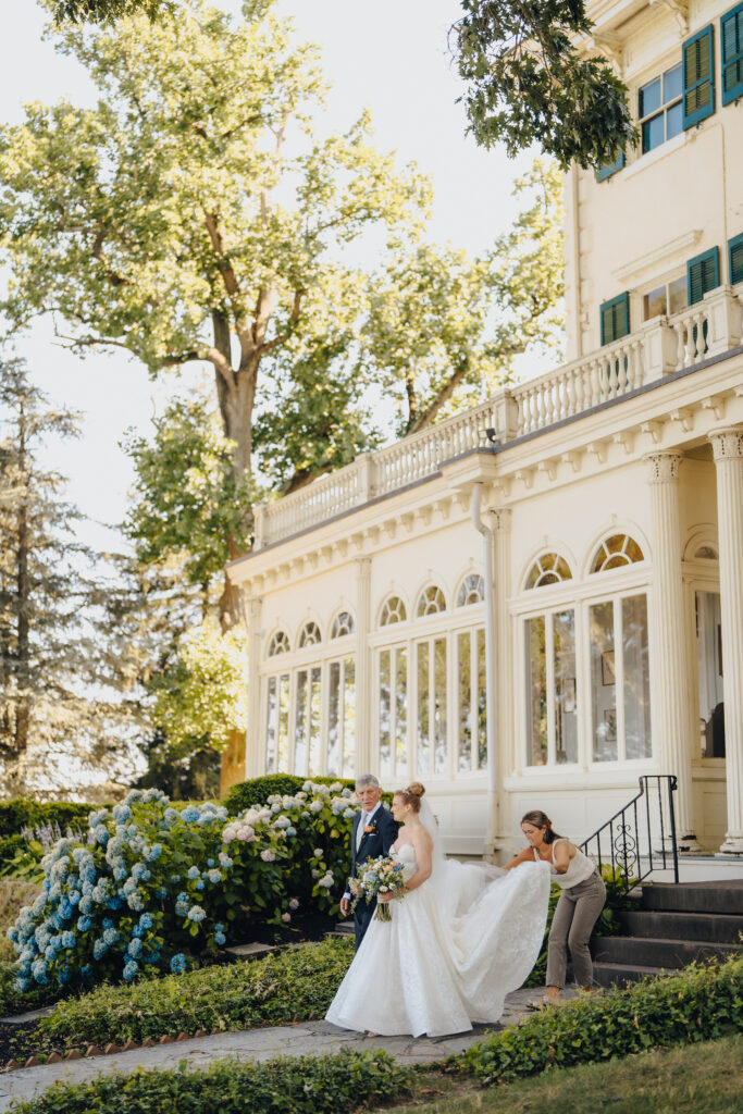 Bride walks with her father at Glen Foerd estate, assisted with her train outside the historic mansion venue in Philadelphia, surrounded by hydrangeas