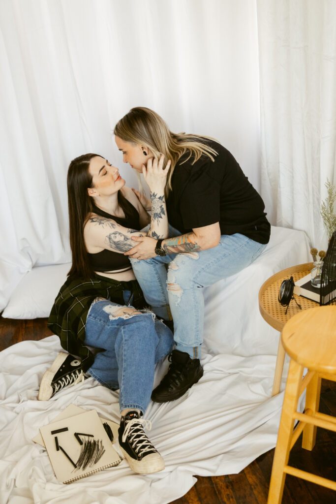 A gay engaged couple; one woman sits on the floor wearing jeans, a black top, and converse, and her partner leans over her wearing a similar outfit.