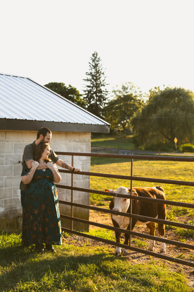 A woman wearing a green dress for her engagement photos, stands while her partner hugs her from behind. The two are outside, looking at one of their cows on their farm!