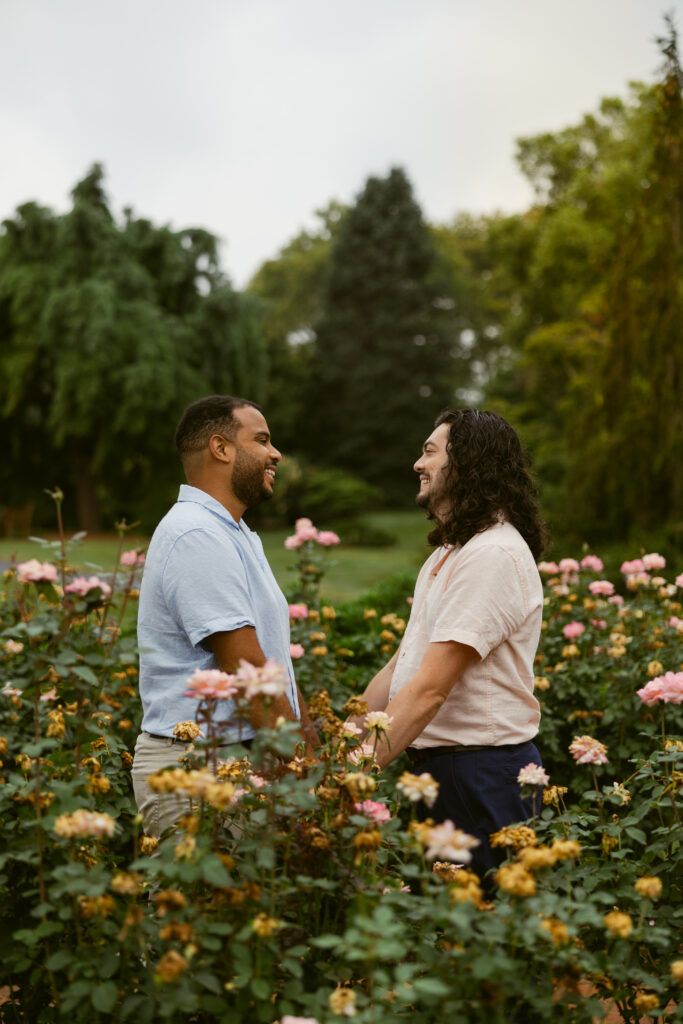 A gay couple standing in roses as they take engagement photos at Hershey Gardens. They're standing facing each other and smiling.