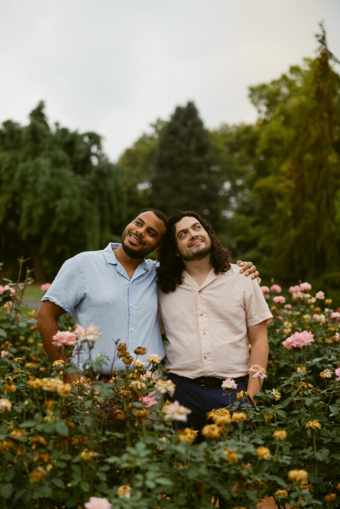 Two men, taking Hershey Gardens engagement photos stand in a rose garden, with the their heads pressed to each other, side-by-side