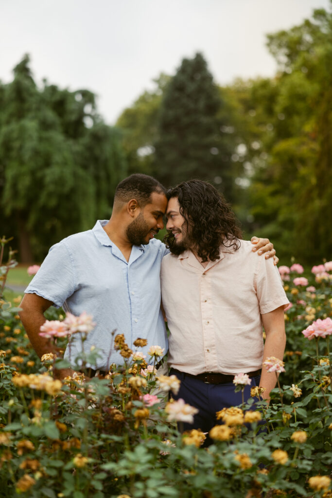 A gay couple standing in roses as they take engagement photos at Hershey Gardens, touching foreheads as their bodies face the camera.