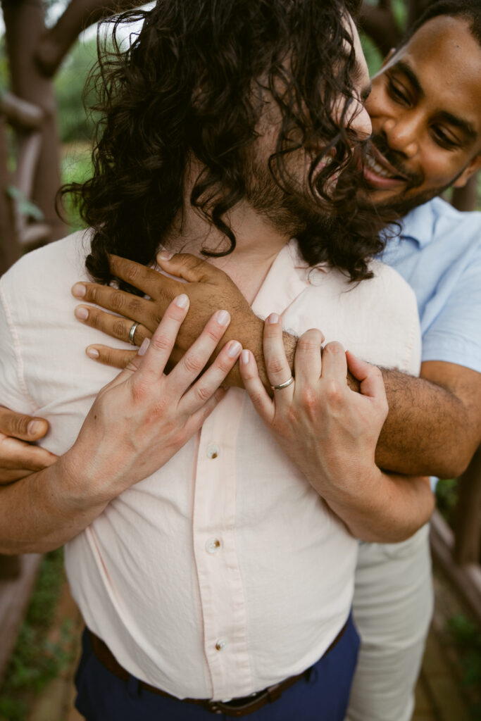 A gay couple poses for engagement photos at Hershey Gardens, one man stands behind his partner with his arm around his chest and the other looks back at his smiling partner