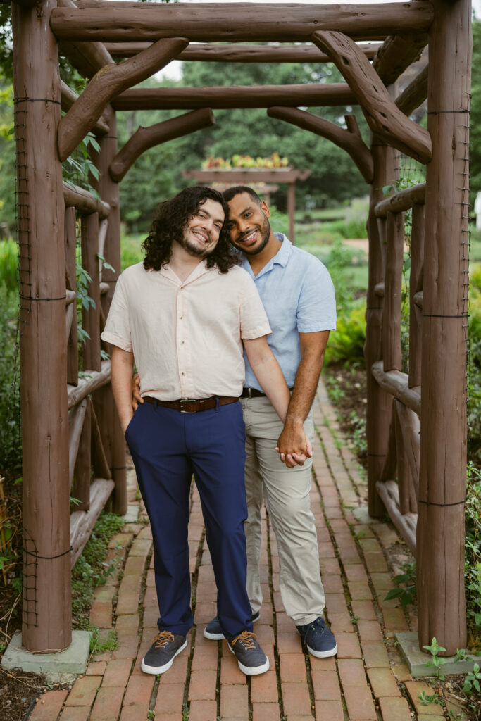 A couple poses beneath a wooden structure, holding hands and hugging as they smile for the camera. One man wears a salmon shirt and blue pants and the other man wears a light blue shirt and khakis 