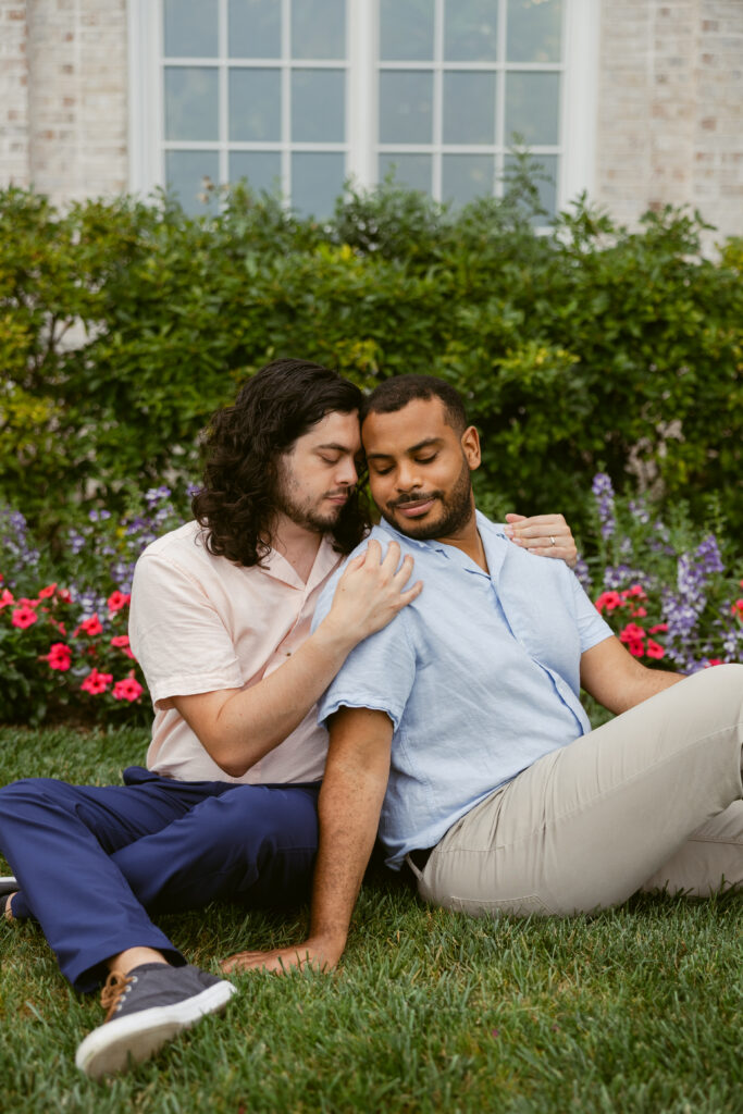 Two engaged men sit on grass, one with his back half turned to his partner who holds his shoulders as he sits behind him