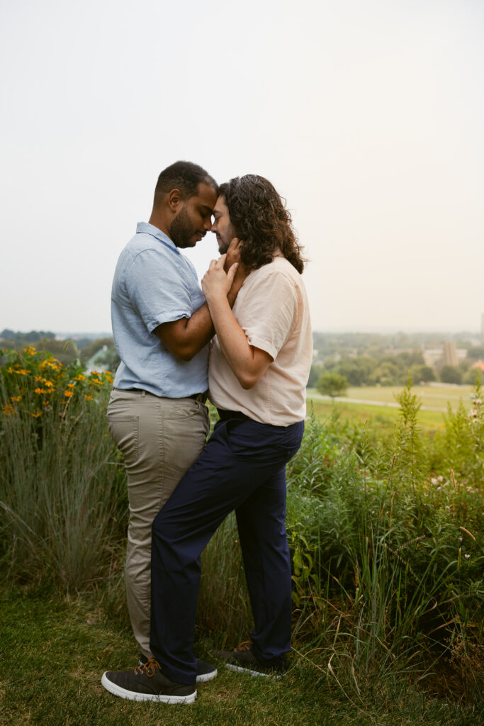 Two men, recently engaged, stand in front of a field of tall, green grass, holding each other's faces with their eyes closed